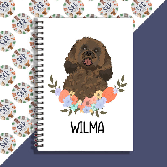 A personalised notebook with an illustration of a cavapoo dog on the cover, surrounded by a floral pattern, with the name 'Wilma' written on the front.