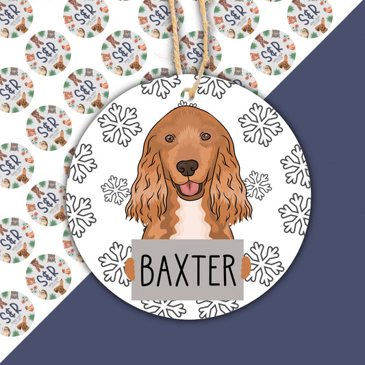 A ceramic Christmas decoration featuring a hand-painted illustration of a Cocker Spaniel dog with snowflakes around it, with the name 'Baxter' printed at the bottom.