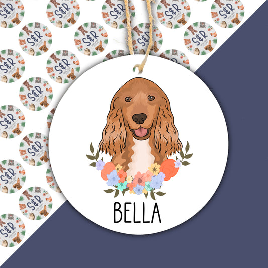 A round ceramic Christmas decoration featuring an illustration of a Cocker Spaniel dog with a floral wreath background, with the name 'Bella' printed at the bottom.