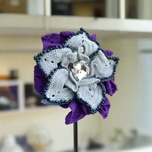 Crocheted flower brooch with purple and grey colors on a stand against a blurred indoor background