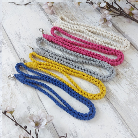 A selection of colourful macrame lanyards laid out on a white wooden surface, available in beige, pink, grey, blue, and yellow colours.