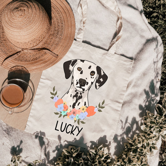 A beige tote bag with a Dalmatian dog illustration and the name 'LUCKY' printed below the dog. The bag is laid out flat with a hat and a glass of water next to it, against a floral background.
