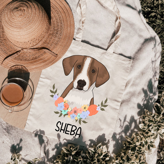A personalised tote bag featuring an illustration of a foxhound dog with a floral wreath and the name 'Sheba' printed on it, placed on a grassy surface with a hat and a cup in the background.