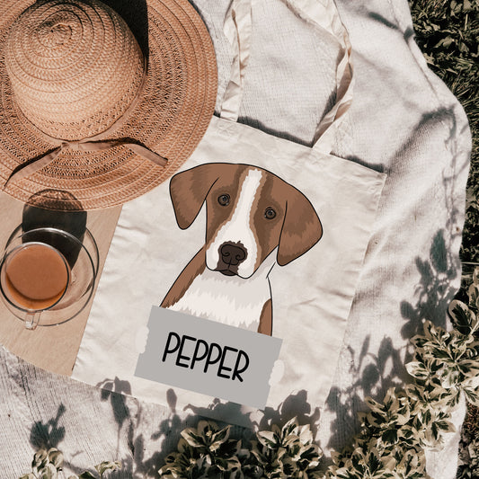 A personalised tote bag with an illustration of a dog breed, named 'PEPPER', on a beige background with a hat and a glass in the background, indicating it is a handmade, customizable gift item.