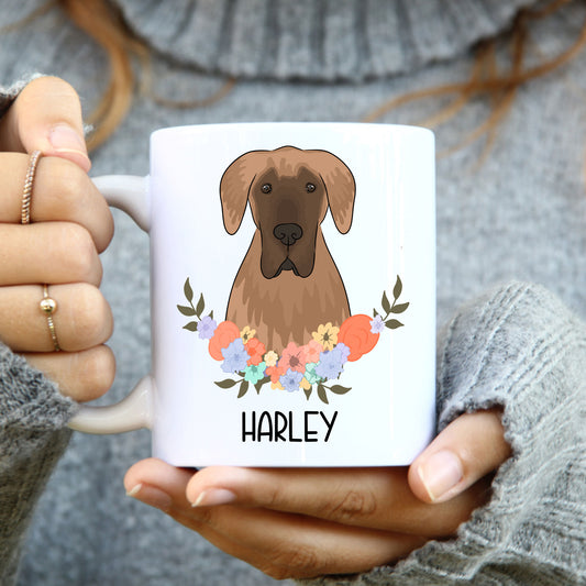A person holding a white coffee mug with a colourful floral design and a graphic of a Great Dane dog in the center.