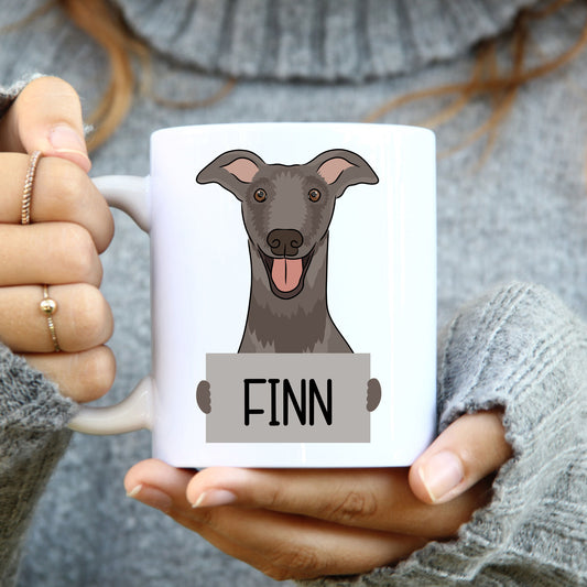 A person holding a white coffee mug with a grayscale illustration of a greyhound dog on it, with the name 'Finn' printed on the mug.