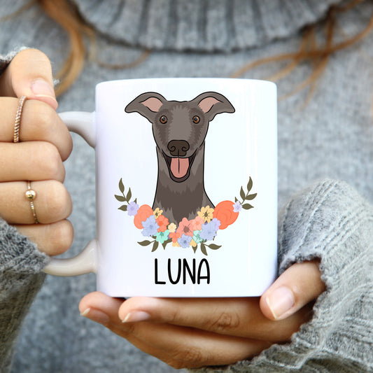 A person holding a white coffee mug with a grayscale illustration of a greyhound dog surrounded by a floral pattern.