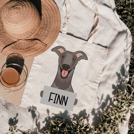 A personalised tote bag featuring a graphic of a greyhound dog with the name 'FINN' printed on it, placed on a grassy surface with a hat and a cup in the background.