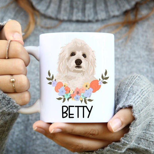 A person holding a white coffee mug with a colourful floral design and an illustration of a Havanese dog breed on the side.
