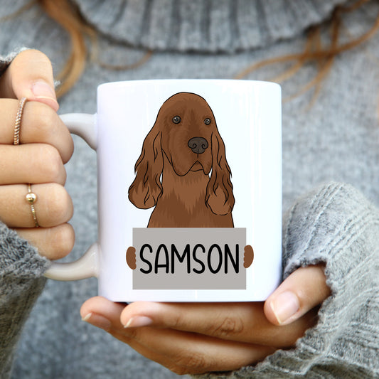A white coffee mug with a colorful illustration of an Irish Setter dog on the side, holding the mug with two hands.