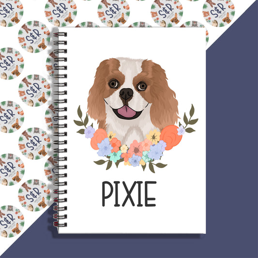 A notebook with a spiral binding featuring a color illustration of a King Charles Spaniel dog on the cover, with the name 'PIXIE' written on the front.