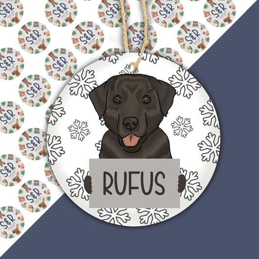 A ceramic Christmas decoration featuring a drawing of a Labrador dog with the name 'Rufus' on a round, white background adorned with snowflakes.