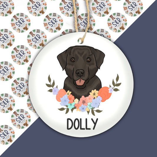 A ceramic Christmas decoration featuring an illustration of a black Labrador dog with a floral wreath around it, with the name 'Dolly' written at the bottom.