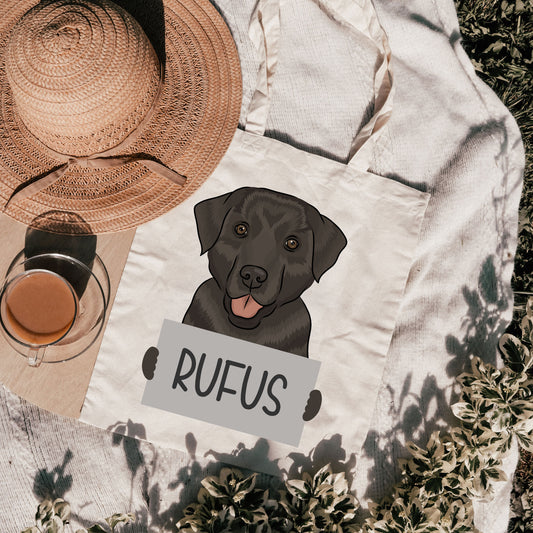 A personalised tote bag with an illustration of a black Labrador dog on the front, with the name 'Rufus' printed below the image. The bag is beige in color and placed on a grassy surface with a straw hat and a cup in the background.