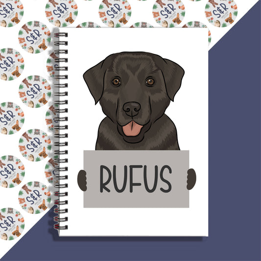A notebook with a spiral binding featuring a graphic of a black Labrador dog on the front cover, with the name 'Rufus' written on the cover.
