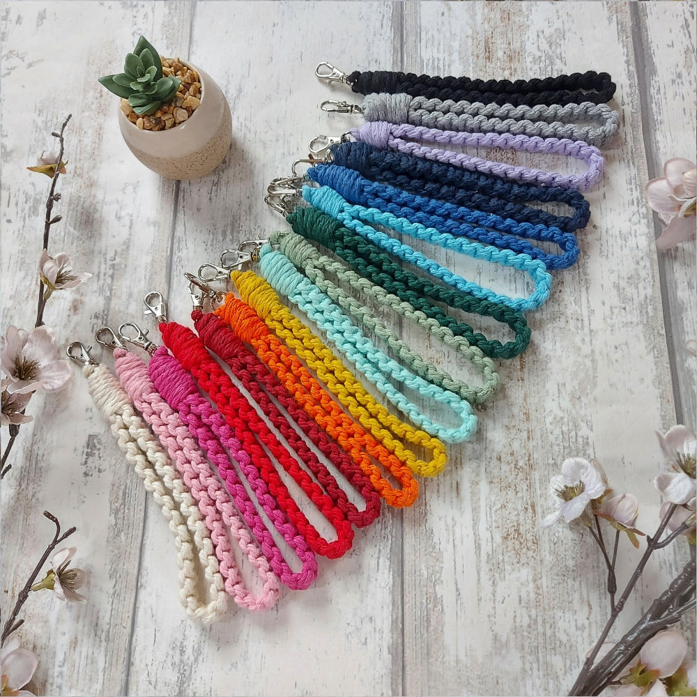 A variety of colourful macrame keyrings displayed on a wooden surface with a small potted plant and floral decoration in the background.