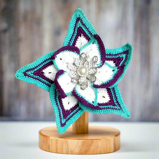 Crocheted flower with purple and teal accents on a wooden stand against a blurred background