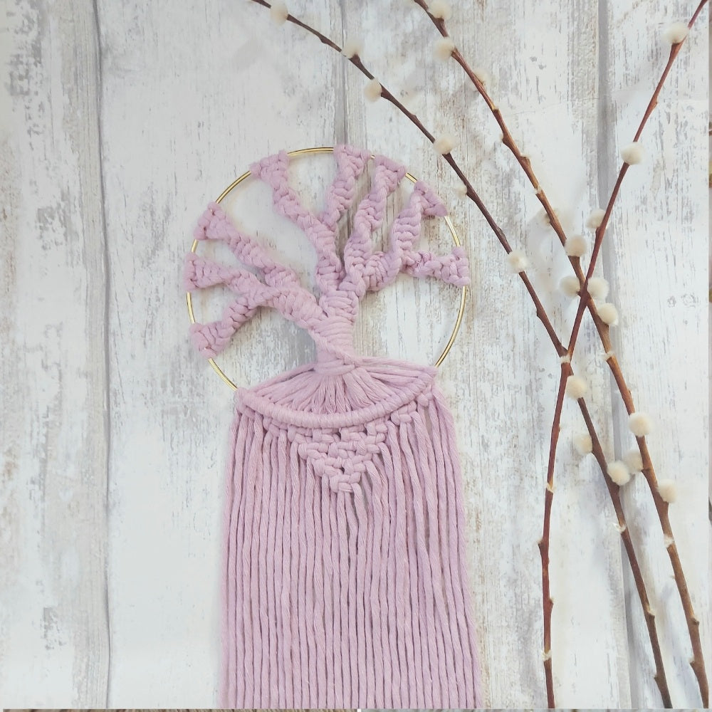 A macrame tree of life wall hanging in pink with a gold-coloured metal hoop and recycled cotton twine, displayed against a white wooden background with branched decorations.