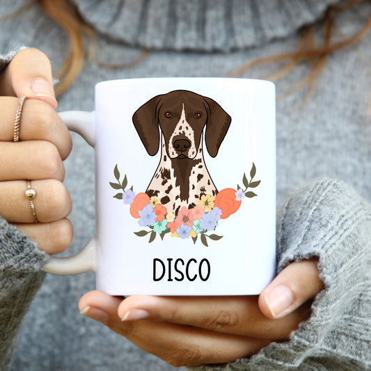A white coffee mug with a colourful illustration of a shorthaired pointer dog accompanied by a floral design, held in a person's hand.