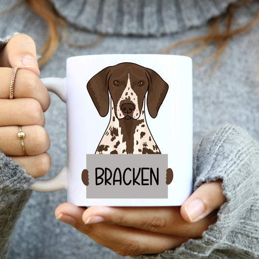 A person holding a white coffee mug with a colourful illustration of a shorthaired pointer dog breed.