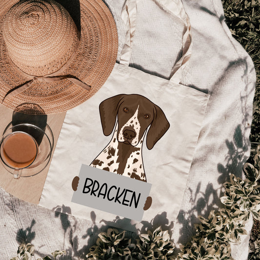 A personalised tote bag with an illustration of a dog breed, named 'Bracken', on a beige background with  a hat and a glass of tea in the background.