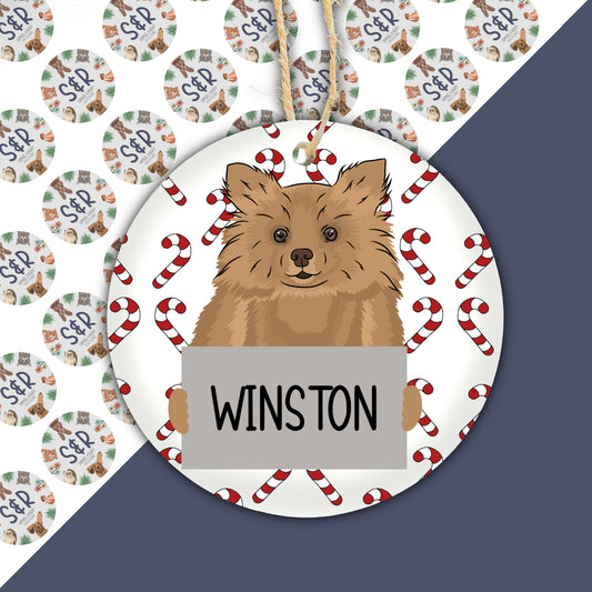 A ceramic Christmas ornament featuring a cartoon illustration of a Pomeranian dog with the text 'Winston' on a tag, surrounded by Christmas-themed patterns.