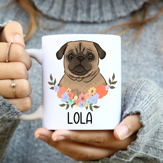 A person holding a white coffee mug with a colourful floral design and a pug dog illustration on it.