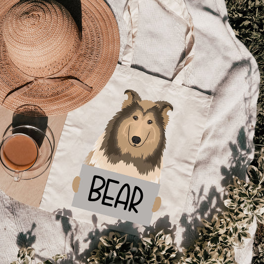 A canvas tote bag with a printed image of a Rough Collie dog breed, with the text 'BEAR' below it, on a outdoor background with a hat and a glass in the background.