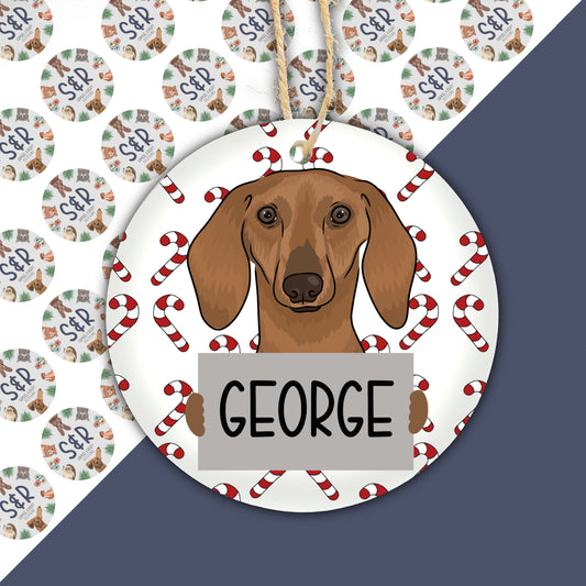 A ceramic Christmas ornament featuring a cartoon-style sausage dog with the name 'George' on a tag, surrounded by Christmas-themed decorations like candy canes.
