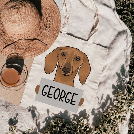 A personalised tote bag with an illustration of a sausage dog and the name 'George' printed on it, placed on a grassy surface with a hat and a cup in the background.