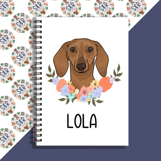 A personalised notebook with a sausage dog illustration on the cover, spiral bound, with floral patterns and the name 'LOLA' displayed on the front.