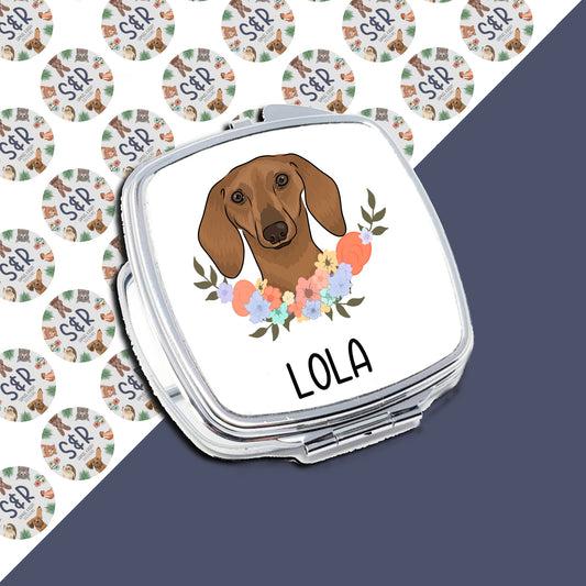 A personalised compact mirror with a sublimated design featuring a cartoon image of a sausage dog (dachshund) with a floral pattern in the background, and the name 'LOLA' at the bottom.