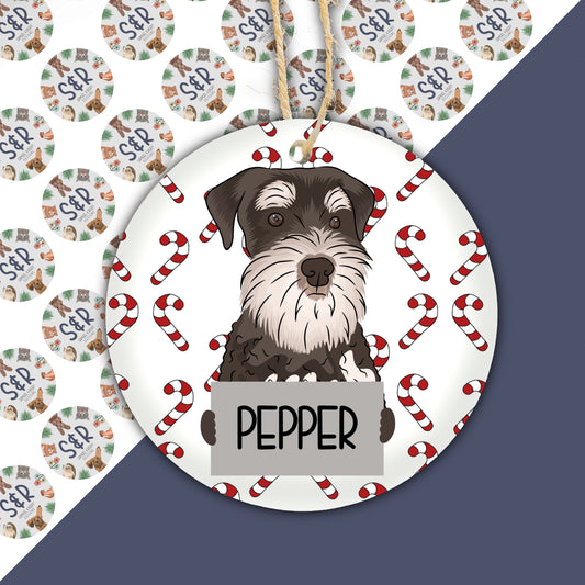 A ceramic Christmas ornament featuring an illustration of a schnauzer dog with the name 'PEPPER' surrounded by Christmas-themed decorations like candy canes.
