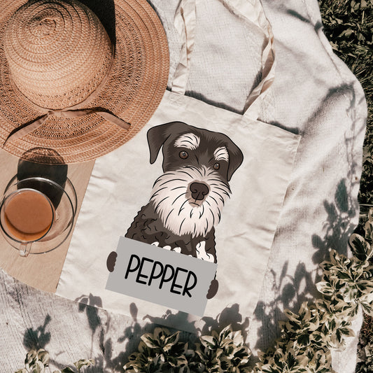 A personalised tote bag with a graphic of a dog on it, placed on a grassy surface with a hat and a cup, with the text 'SCHNAUZER' on the front.