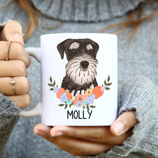 A person holding a white coffee mug with a colourful floral design and an illustration of a Schnauzer dog on the side.