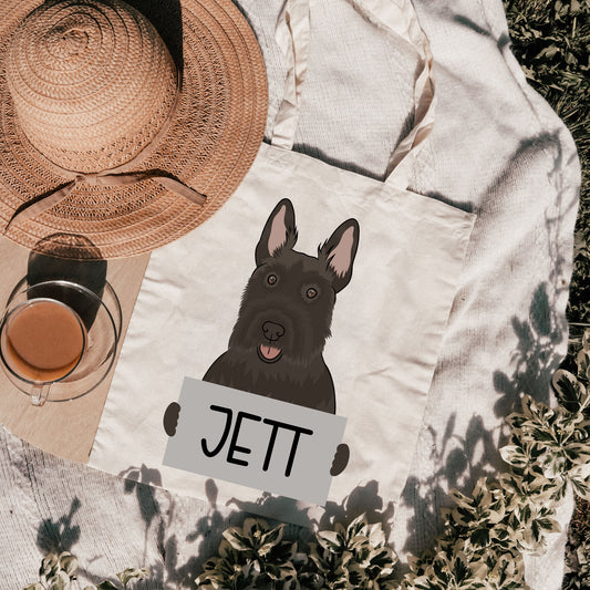 A personalised tote bag with an illustration of a Scottish Terrier dog and the name 'Jett' printed on it, placed on a grassy surface with a hat and a cup in the background.