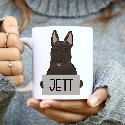 A person holding a white coffee mug with an illustration of a Scottish Terrier dog on it, with the name 'JET' written on the mug.