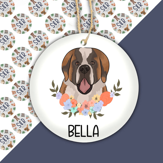 A ceramic Christmas decoration featuring a colourful illustration of a Saint Bernard dog with a floral wreath background, with the name 'Bella' printed at the bottom.