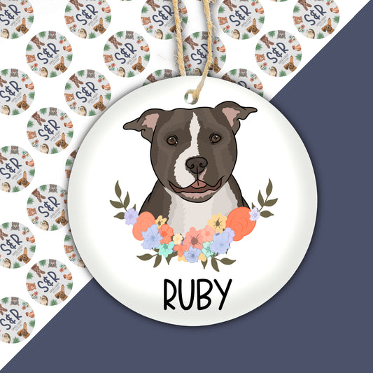 A round ceramic Christmas decoration featuring an illustration of a Staffordshire Bull Terrier dog with a floral wreath background, with the name 'Ruby' underneath.