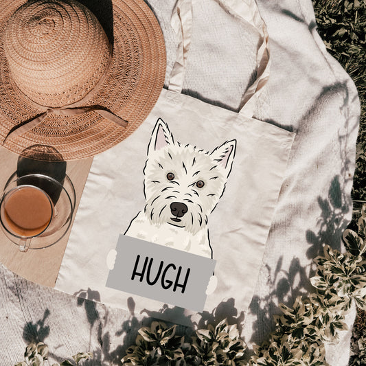 A white personalized tote bag with an illustration of a Westie dog on it, placed on a grassy surface with a hat and a cup, with the name 'HUGH' printed on the front.