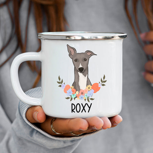 A white enamel mug with a illustration of a whippet dog surrounded by a floral wreath, with the name 'ROXY' inscribed on the mug.