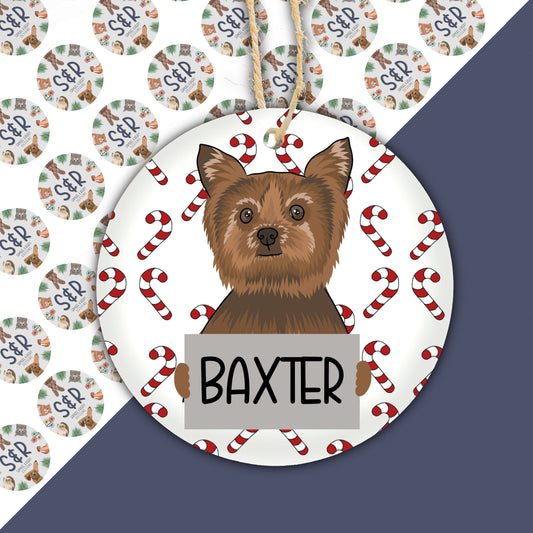 A ceramic Christmas ornament featuring an illustration of a Yorkie dog surrounded by Christmas-themed decorations, with the name 'Baxter' printed at the bottom.